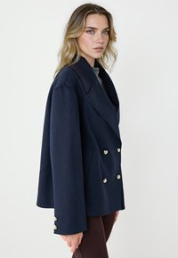 Blazer doppiopetto blu navy con ampio rever, bottoni color oro e tasche laterali, caratterizzato da una silhouette strutturata e una texture liscia.