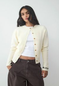 Cremfarbener Cardigan aus weichem Material mit Rundhalsausschnitt und Holzknöpfen. Über einem weißen Oberteil getragen und kombiniert mit dunklen Hosen.