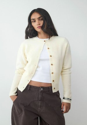 Crème-kleurige cardigan gemaakt van zacht materiaal met een ronde hals en houten knopen. Draagt over een witte top en gecombineerd met donkere broek.