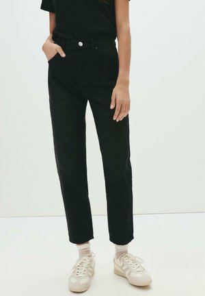 Jean noir taille haute avec jambe droite, poches avant et fermeture à bouton. Porté avec des baskets blanches et des chaussettes crew.