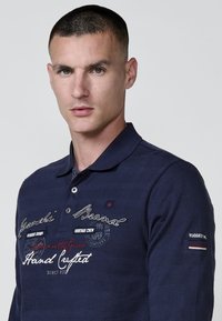 Jūras zils polo krekls, kas izgatavots no teksturēta auduma, ar apkakli, trim pogām un izšuvumiem ar "Rugged Brand" un citiem teksta elementiem.