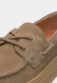 Καφέ παπούτσι τύπου boat από σουέντ με δερμάτινα κορδόνια που περνούν μέσα από μεταλλικά γαντζάκια και ραφές στη σόλα.