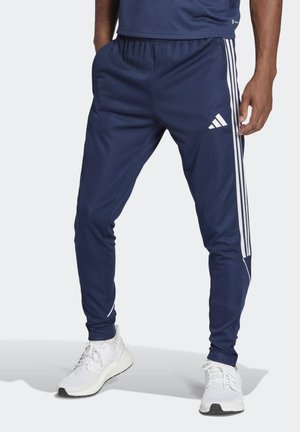 Mężczyzna ubrany w granatowe spodnie dresowe Adidas z białymi paskami i logo, w zestawieniu z białymi butami sportowymi, stojący na jednolitym tle.