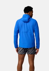 Giacca a cappuccio blu con tessuto liscio, maniche aderenti e orlo curvo. Abbinata a pantaloni corti grigi con motivo a strisce diagonali.