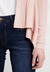 Person in dunkelblauen Jeans und einer hellrosa gerippten Strickjacke mit asymmetrischem Saum, die Hand entspannt an der Seite.
