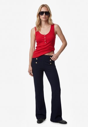 Frau mit schwarzen Sonnenbrillen, einem ärmellosen roten Ripptop mit Knöpfen, marineblauen Hosen mit goldenen Knöpfen und schwarzen Schuhen, stehend vor weißem Hintergrund.