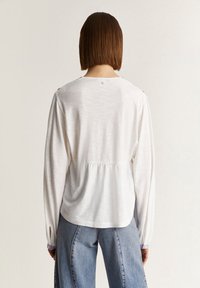 Valkoinen pitkähiha blouse, jossa on kootut vyötärö ja napit olkapäillä, sekä teksturoitu kangas. Yhdistetty löysiin sinisiin farkkuihin.