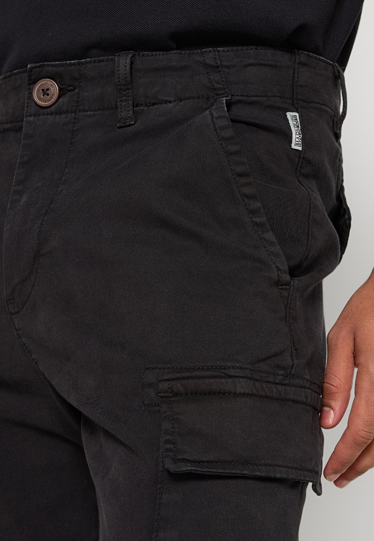 Primer plano de una persona que lleva pantalones cargo negros con un bolsillo con tapa y un botón visible, con la mano descansando al lado del muslo.