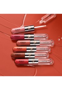 Zes KIKO Milano Unlimited Double Touch lipgloss-tubes in verschillende tinten, gerangschikt op een gladde koraalkleurige achtergrond met glanzende vegen.