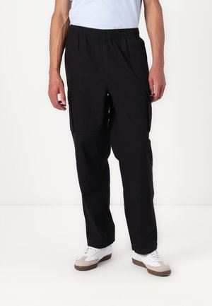 Black cargo pants made of fabric with an elastic waist. Features side pockets and a straight-leg design. Paired with white sneakers.