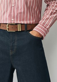 Camisa a rayas rojas y blancas combinada con jeans de mezclilla azul oscuro. Cinturón marrón con hebilla rectangular de latón. Diseño clásico y corte ajustado.