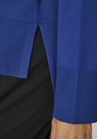 Maglione lavorato a maglia blu con texture a coste, con spacco laterale e polsini a contrasto. Tessuto nero visibile sotto.