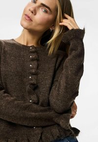 Brun stickad tröja med rund halsringning, volang framtill och dekorativa knappar. Har långa ärmar med volangvidder och textil med struktur.