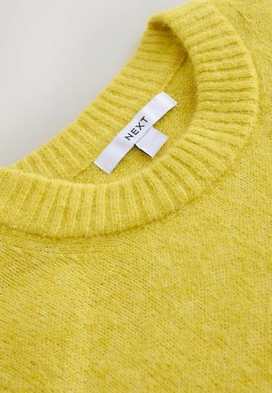 Dzeltens adīts džemperis ar ribotu apaļu kakla izgriezumu un mīkstu tekstūru. Ožzīme ar uzrakstu "NEXT". Tuva kadra attēls, kas izceļ krāsu un materiālu.