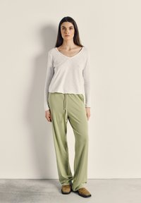 Ženska stoji ob bež steni, oblečena v belo dolgimi rokavi z V-izrezom, svetlo zelene hlače z vrvico in rjave čevlje.