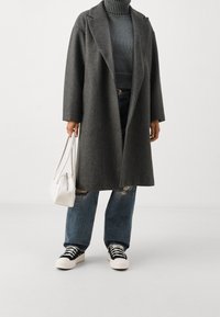 Grå ullkappa med klockad krage, ihop med en ribbad mörk tröja, slitna blåjeans och svarta och vita sneakers. Vit handväska.