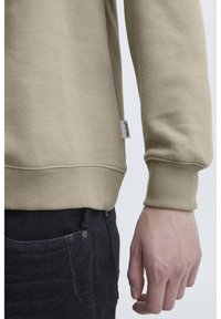 Sudadera beige con puños y dobladillo acanalados, hecha de una tela suave. Presenta una pequeña etiqueta de la marca en un lado y se combina con jeans oscuros.