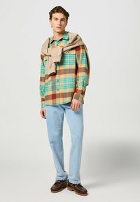 Camisa de cuadros en azul, amarillo y marrón, con mangas largas y botones; suéter beige sobre los hombros; jeans azul claro; zapatos marrones.