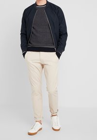 Giubbotto bomber blu navy, maglione grigio a maglia, chinos beige e sneakers bianche con suole marroni. Il giubbotto presenta una chiusura con zip e polsini a coste.