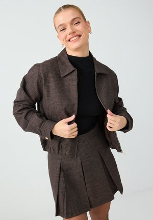 Un mannequin souriant tient ouverte une veste marron à carreaux sur un haut noir et une jupe plissée assortie, devant un fond de studio uni.