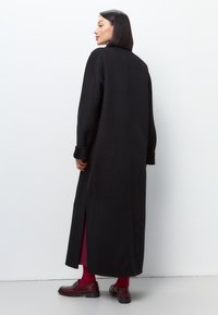 Cămașă de lână neagră, cu croială dreaptă, mâneci lungi și fante laterale. Purtați peste o rochie bordeaux, asortată cu loaferi lucioși negri.