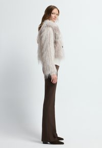Chaqueta de piel sintética en color beige claro con flecos texturizados, llevada sobre un suéter blanco y pantalones acampanados marrones, completada con zapatos oscuros de punta.