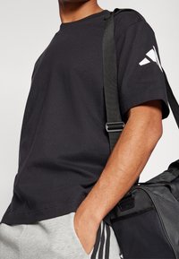 Černé bavlněné tričko s krátkými rukávy, spárované s světle šedými tepláky. Černá taška na rameno s texturovaným povrchem a viditelným sponou.