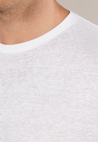 Biała koszulka typu crew-neck z teksturowanej, lekkiej tkaniny. Zbliżenie ukazuje gładki dekolt i subtelny wzór na materiale.