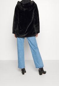 Cappotto di pelliccia sintetica nera con cappuccio, taglio oversize, maniche lunghe e una consistenza morbida, abbinato a jeans a gamba larga blu chiaro e stivali neri alla caviglia.