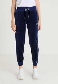 Navy modré velurové tepláky s bílými bočními pruhy, elastickým pasem a šňůrkou. Logo Nike v bílé barvě na levém boku.
