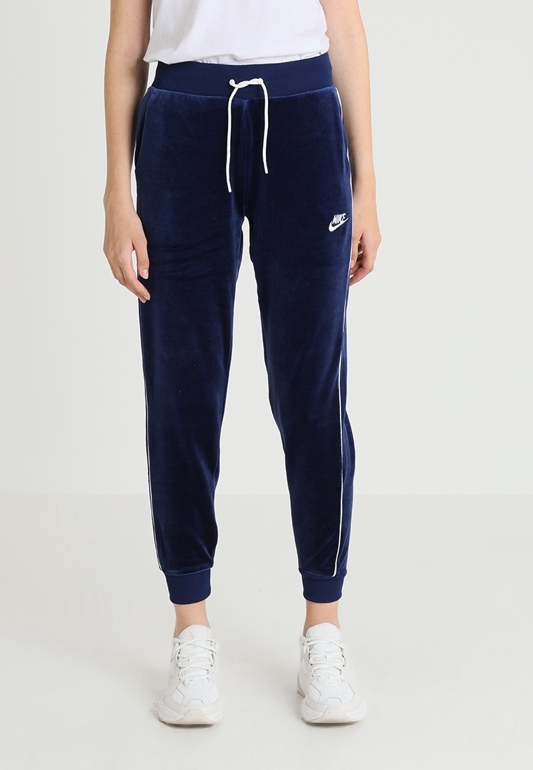 Navy modré velurové tepláky s bílými bočními pruhy, elastickým pasem a šňůrkou. Logo Nike v bílé barvě na levém boku.