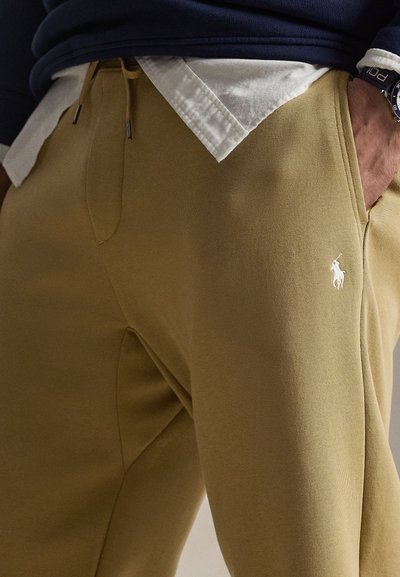 Man draagt beige joggingbroek met wit geborduurd Polo-logo, wit overhemd deels ingestopt, marineblauwe trui en horloge aan linkerpols.