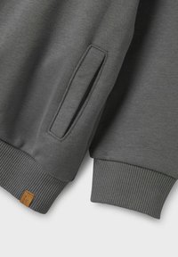 Grå sweatshirttyg som visar ribbade muddar, en sidoficköppning och en läderetikett på fållen. Slät struktur med en enhetlig färg.