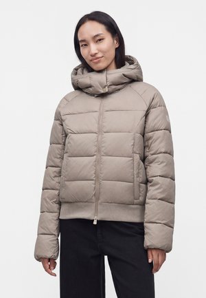 Jeune femme portant une veste matelassée beige à capuche et un pantalon noir, debout devant un fond blanc uni.