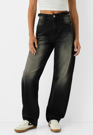 Personne portant un jean large noir délavé à taille haute, un crop top blanc et des baskets blanches avec des accents noirs, debout devant un fond blanc.