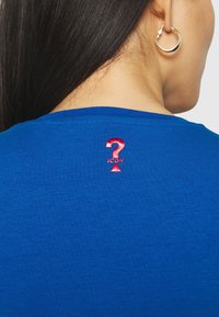 Sininen puuvillainen t-paita, jossa on pyöreä kaula-aukko, ja jossa on takana punainen brodeerattu '?' ja sen alla sana 'IKONI'.