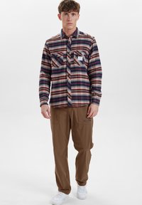 Camisa de franela a cuadros en azul marino, rojo y crema; dos bolsillos en el pecho; combinada con pantalones marrones; zapatillas blancas; fondo liso.