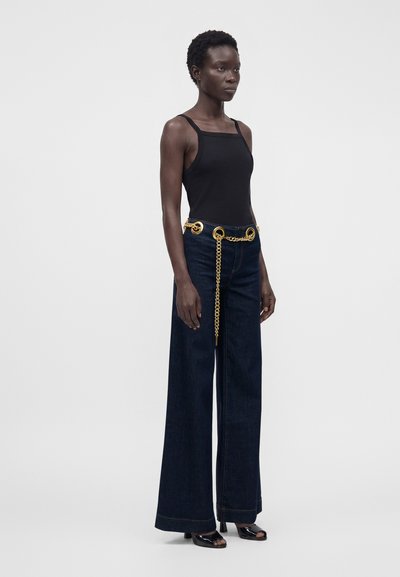 Kvinde stående iført sort ærmeløs top, mørke vidde jeans med gyldent kædebælte og sorte åbne hæle mod en ensfarvet baggrund.