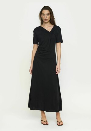 Moteris dėvi juodą, trumpomis rankovėmis, iki čiurnos ilgio suknelę su V formos iškirpte ir šiek tiek surinkimu, stovinti priešais paprastą baltą foną.