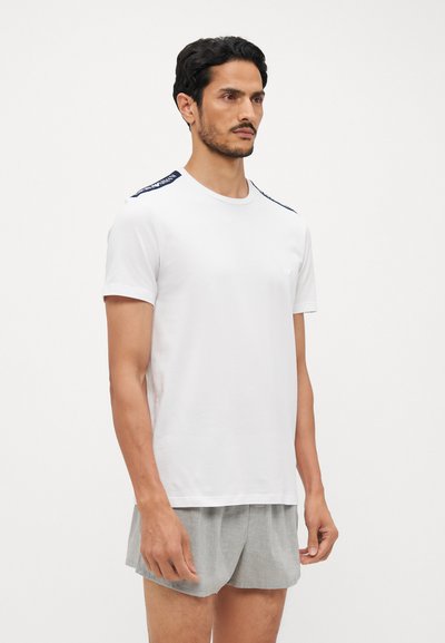 T-shirt bianco in cotone con maniche corte, scollatura rotonda e dettagli blu navy sulle spalle. Il modello indossa pantaloni corti grigi chiari.
