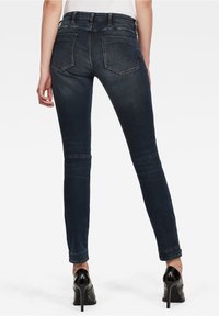 Jeans ajustados de color azul oscuro con bolsillos traseros, que presentan una textura desvaída y una cintura de altura media. Combinados con zapatos de tacón alto negros.