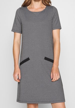 Vrouw draagt een zwart-witte jurk met korte mouwen, knielengte en twee zwarte diagonale zakdetails aan de voorkant.