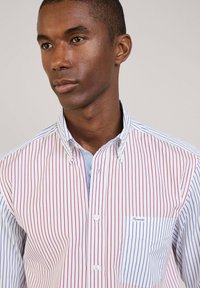 Camicia a bottone con strisce verticali rosse e blu su uno sfondo bianco. Ha un colletto e una piccola tasca sul petto con il marchio.