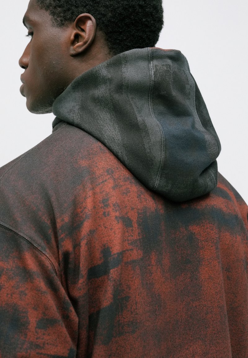 Ung mand, der står med ansigtet til venstre, iført en mørkerød og sort mønstret hættetrøje med hætten nede, mod en simpel lys baggrund.