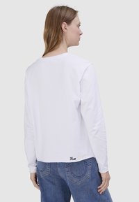 Weißes Langarmshirt mit rundem Ausschnitt. Verfügt über einen leicht verkürzten Schnitt und eine in Schwarz und Gold gestickte Markenschrift am Saum.