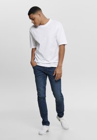 White cotton t-shirt with short sleeves paired with dark blue jeans. Model stands in a relaxed pose, wearing white sneakers.