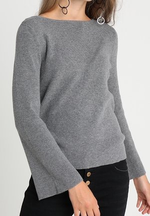 Grå strikket sweater med bådudskæring, lange klokkeærmer og en afslappet pasform. Et par knapper synlige på sorte bukser.