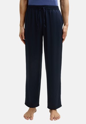 Man in marineblauwe pyjamabroek met trekkoord, blootsvoets staand tegen een effen lichte achtergrond.