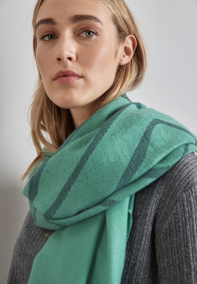 Écharpe verte surdimensionnée avec des rayures foncées, fabriquée en tissu doux et texturé, enroulée autour des épaules, associée à un pull gris à côtes.