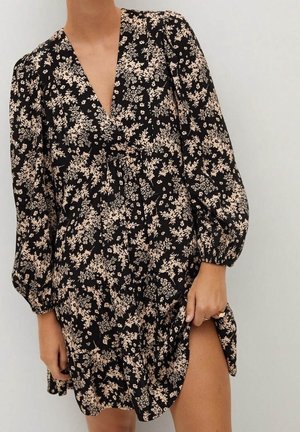 Frau trägt ein schwarzes, langärmeliges Kleid mit beigen Blumen, tiefem V-Ausschnitt und seitlichem Schlitz, steht vor einem schlichten hellen Hintergrund.